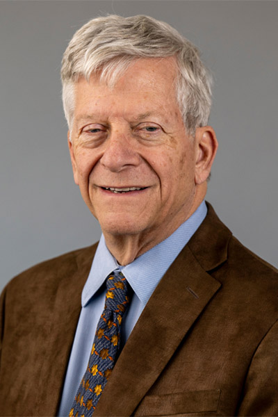 Portrait of a man with short hair, brown blazer, light blue shirt and a blue, gold and orange geometric tie.